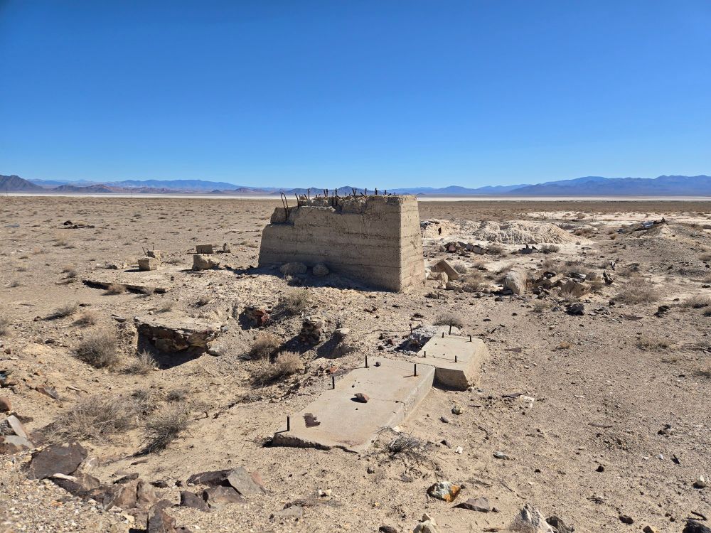 A wide flat valley under a clear blue sky. In the center is the ruins of some mining operation. A weathered pit is to the left with an unseen rock wall on one side. Towering in the middle is a large concrete footing type structure with numerous bolts sticking out the top. In the distance is a patch of white material that shows signs of being dug around in. Surrounding the area is debris of concrete, metal, wood, and small bits of colored glass fragments.