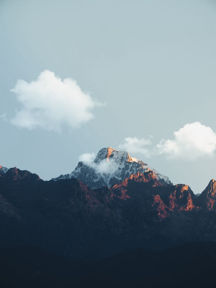 A mountain peak in the area of sikkim, a part of India. The sunlight hits the snow covered mountaintop with warm light and some clouds are stuck there as well. 