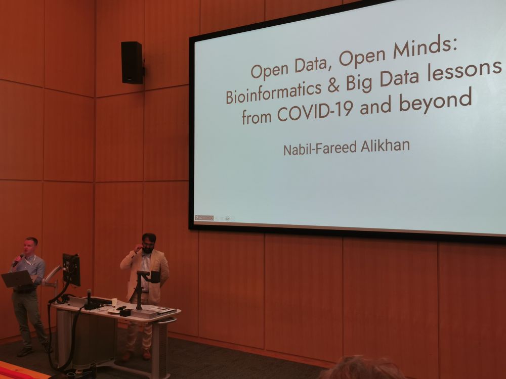 Prof Nik Morton hosts Dr Nabil-Fareed Alikhan in front of a lectern with title slide "Open data, open minds: bioinformatics and big data lessons from COVID-19 and beyond"