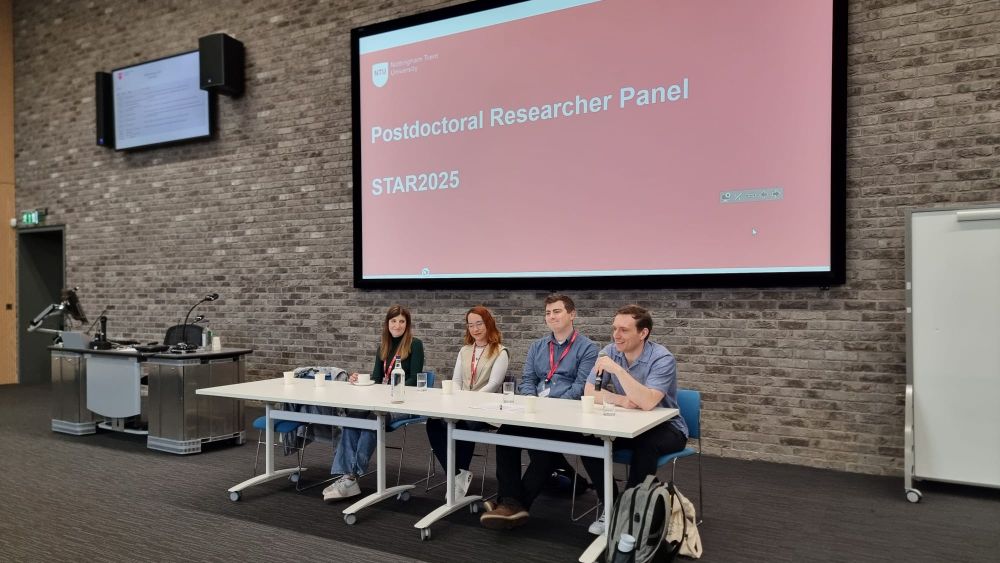 Four postdocs (two women two men) sat at a panel desk in front of a slide saying "postdoctoral researcher panel"