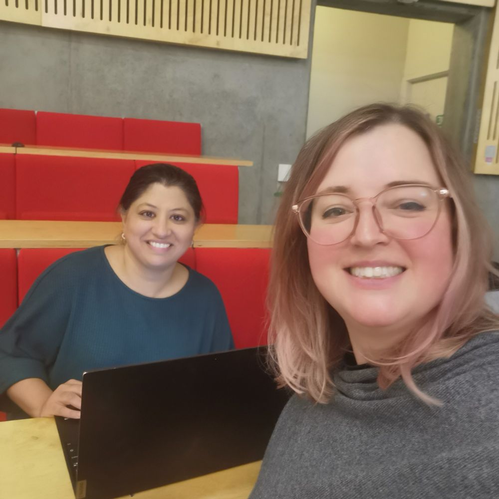 Preeti and Rebecca smiling in an empty lecture theatre. Preeti is sat in front of a laptop