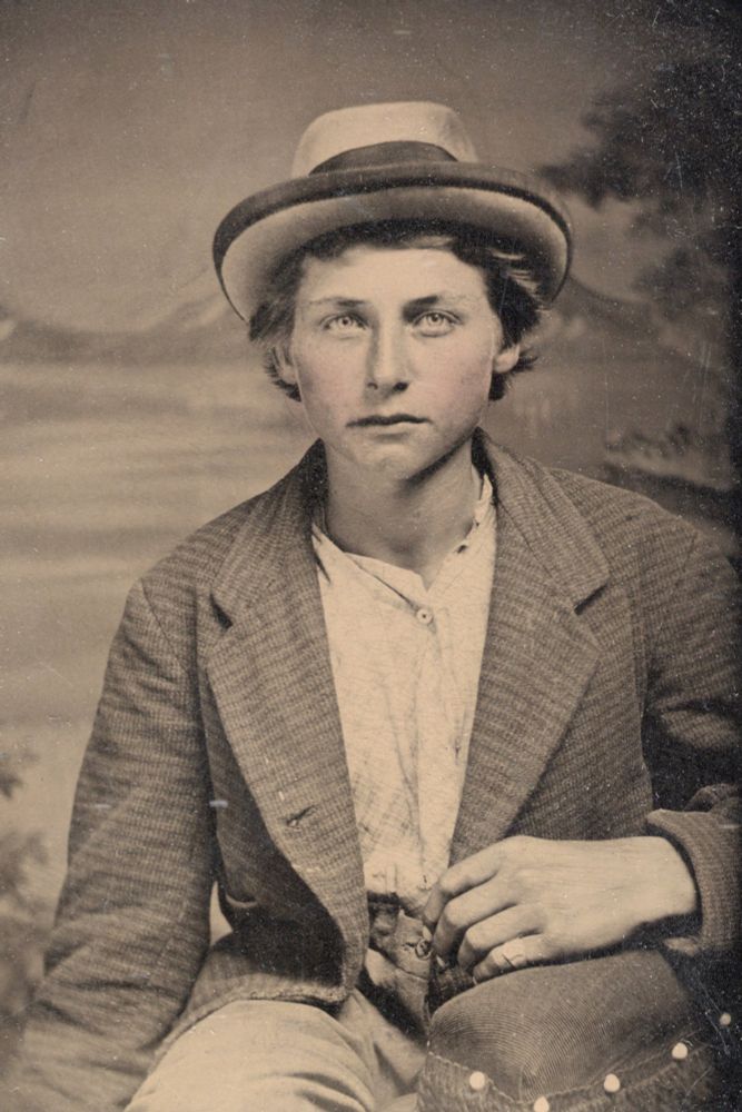 Black and white vintage photo of a white teenage boy with a heart-shaped face, brown hair, and piercingly light eyes, wearing a brimmed hat and a jacket over a white button-up shirt.