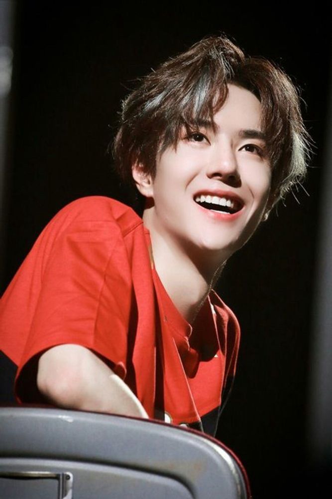 Wang Yibo smiling widely, with his mouth slightly open and the left side of his smile quirking even more sideways in amusement. His eyes are also smiling. His hair is very full and floofy, with silver-white highlights. He has on a red tshirt and is sitting in a gray meta chair, turning around toward the side of the chair, looking up and off-camera.