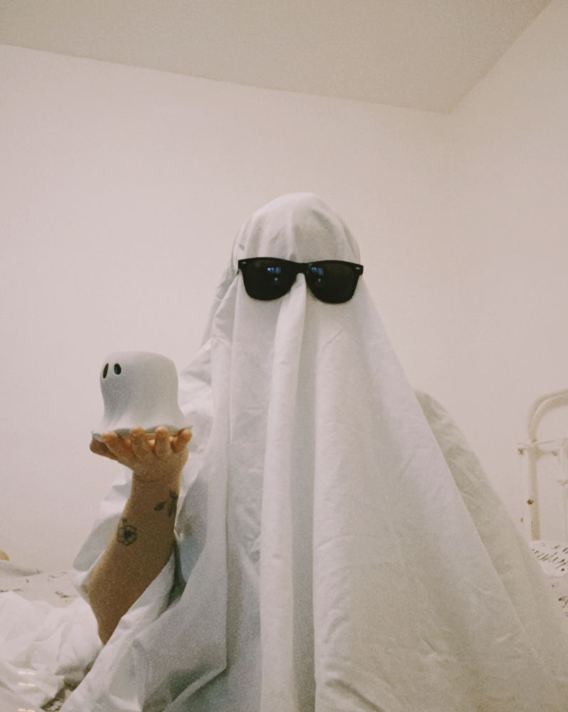 toni covered in a white sheet with black sunglasses on and holding out a small white ghost figuring in their left hand