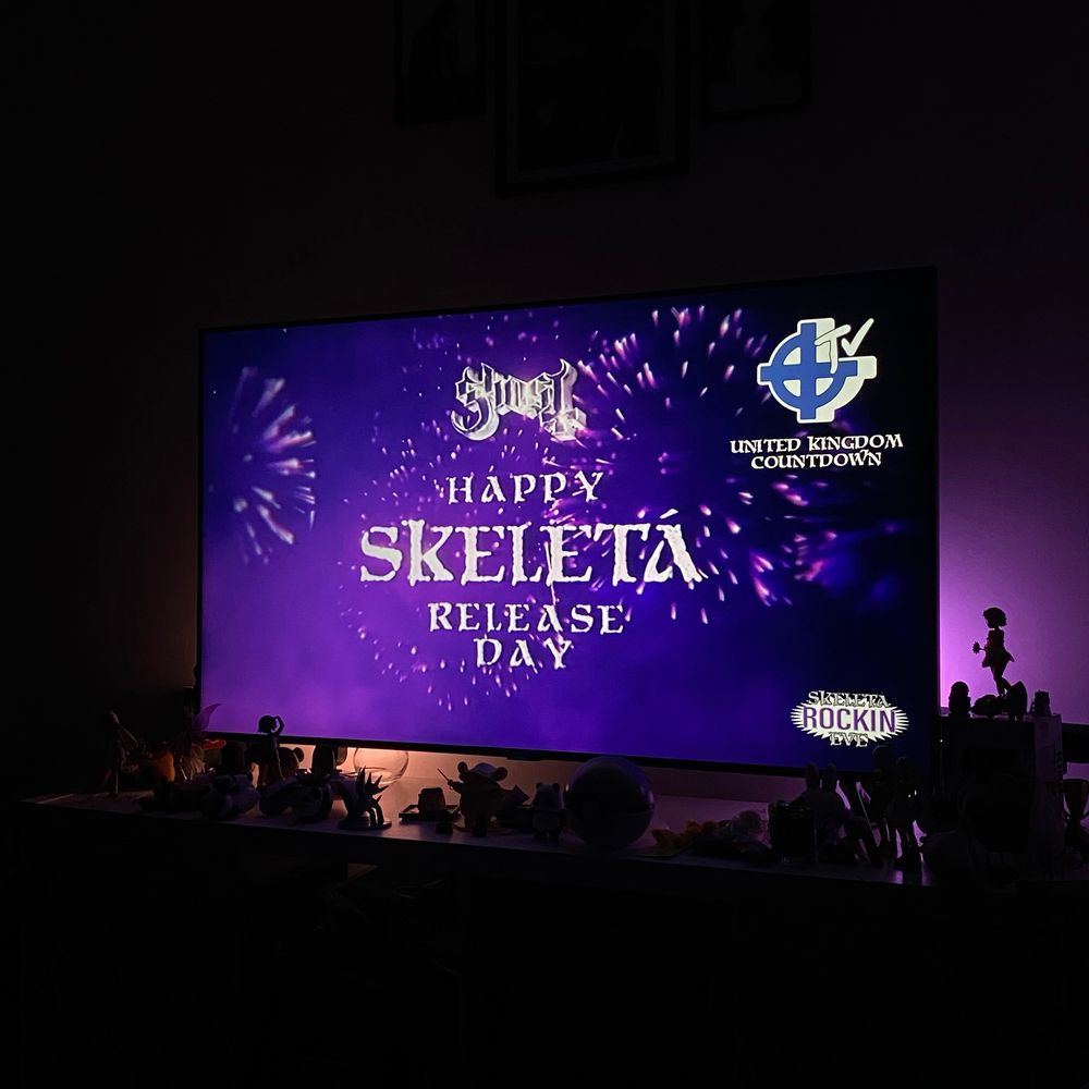 A TV screen showing the UK countdown live stream for Ghost’s new album Skeleta. The screen is paused on the “Happy Skeleta Release Day” text with a purple background with fireworks.