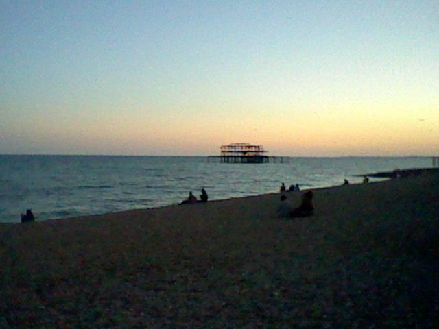 Brighton West Pier at sunset, sillhoutted people are sat on the pebbled beach