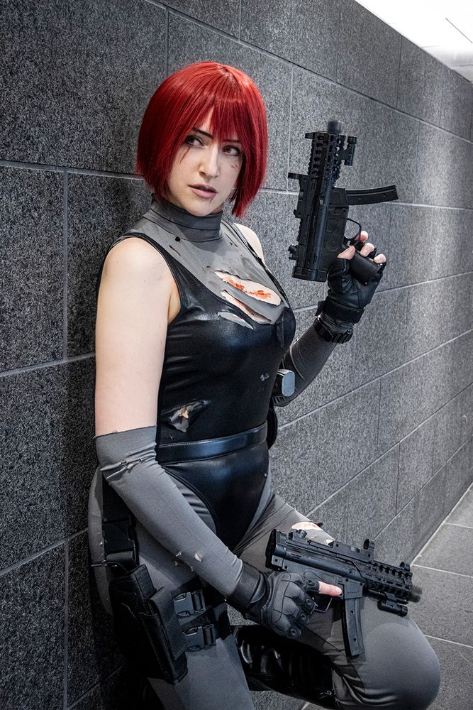 Sheenah is cosplaying as Regina from the video game Dino Crisis, wearing her default outfit with some battle damage and fake blood. She's leaning against a wall, holding two fake machine guns and looking sideways.
