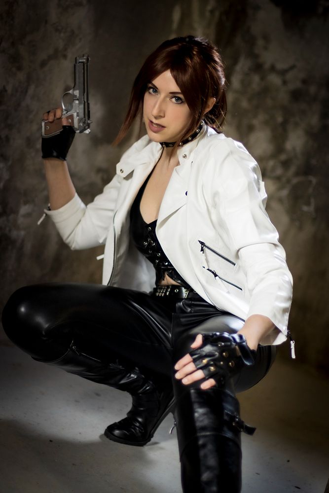 Sheenah is cosplaying as Claire Redfield from the video game Resident Evil: The Darkside Chronicles, wearing her black and white biker bonus outfit. She's crouching and holding a fake gun, looking at the camera.