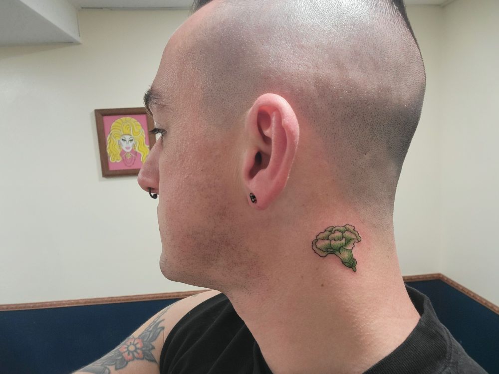 Me, white late thirties person with a black sleeveless shirt and black stud earrings and septum ring, am turned away from the camera in a green and white tattoo studio with a traditional tattoo style piece of art depicting Trixie Mattel in the back ground, showing off my new 1 inch neck tattoo of a green carnation