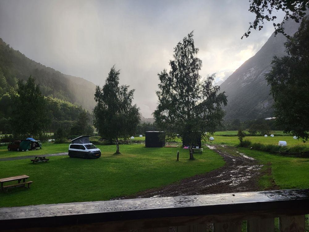 Blick von einer Terrasse auf eine Wiese. Dort stehen ein paar Bäume und wohnmobile. Zwischen den Bergen im Hintergrund kommt gerade eine Regenschauer runter.

View from a terrace onto a meadow. There are a few trees and campers. A rain shower is falling between the mountains in the background.
