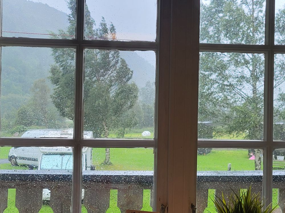 Hinter einem Fenster sind Bume die sich leicht durch den Sturm biegen. Diese stehen sowie auch zwei Wohnmobile auf einer grünen Wiese. Im Hintergrund ist auf der linken Seite ein Berg.

Behind a window are trees, slightly bent by the storm. These, along with two mobile homes, are standing on a green meadow. In the background, on the left, is a mountain.