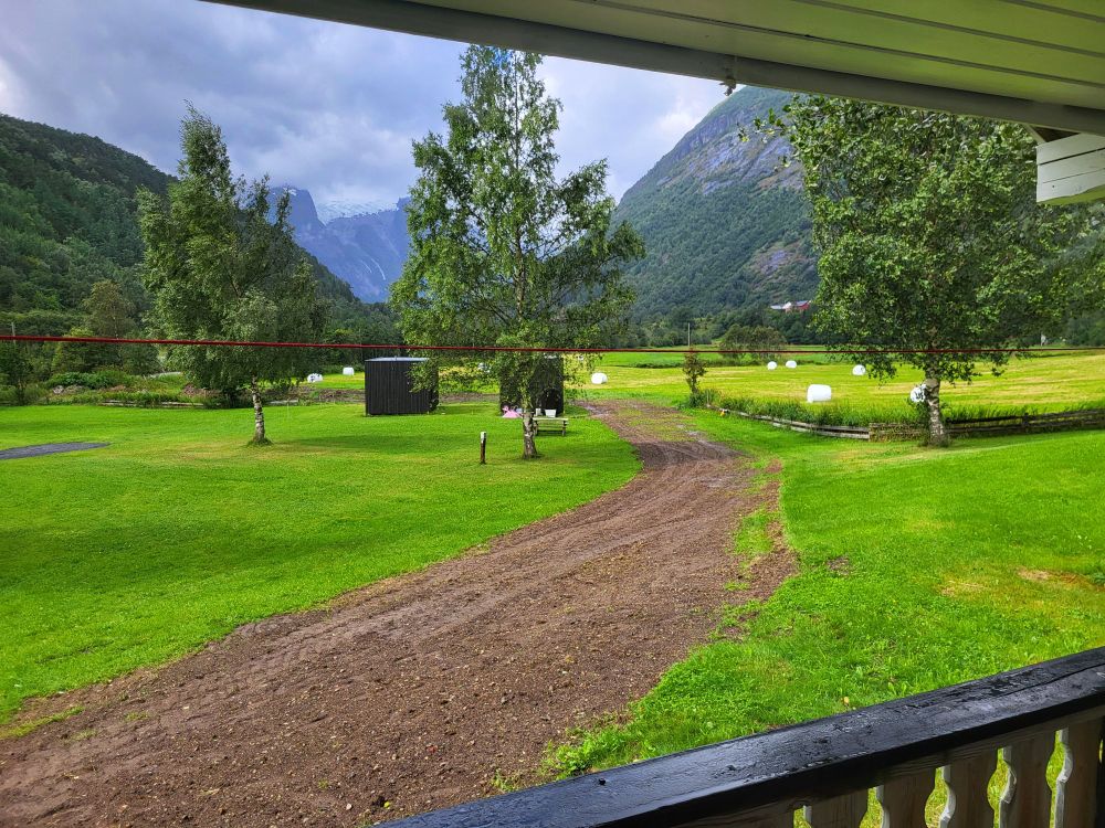 Blick von einer Terrasse auf eine Wiese mit einem Weg de kompltte mit Erde bedeckt ist. Auf der Wiese stehen drei Bäume. Im Hintergrund befinden sich Berge.

View from a terrace onto a meadow with a path completely covered with earth. There are three trees in the meadow. Mountains are in the background.