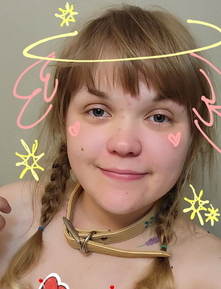 A selfie of me, Cupid. I'm smiling at the camera with medium blonde hair in twin braids with a tan collar around my neck. I have a halo, wings, and stars drawn around me in a crayon style and my logo drawn by freakarchives in the bottom left corner. 