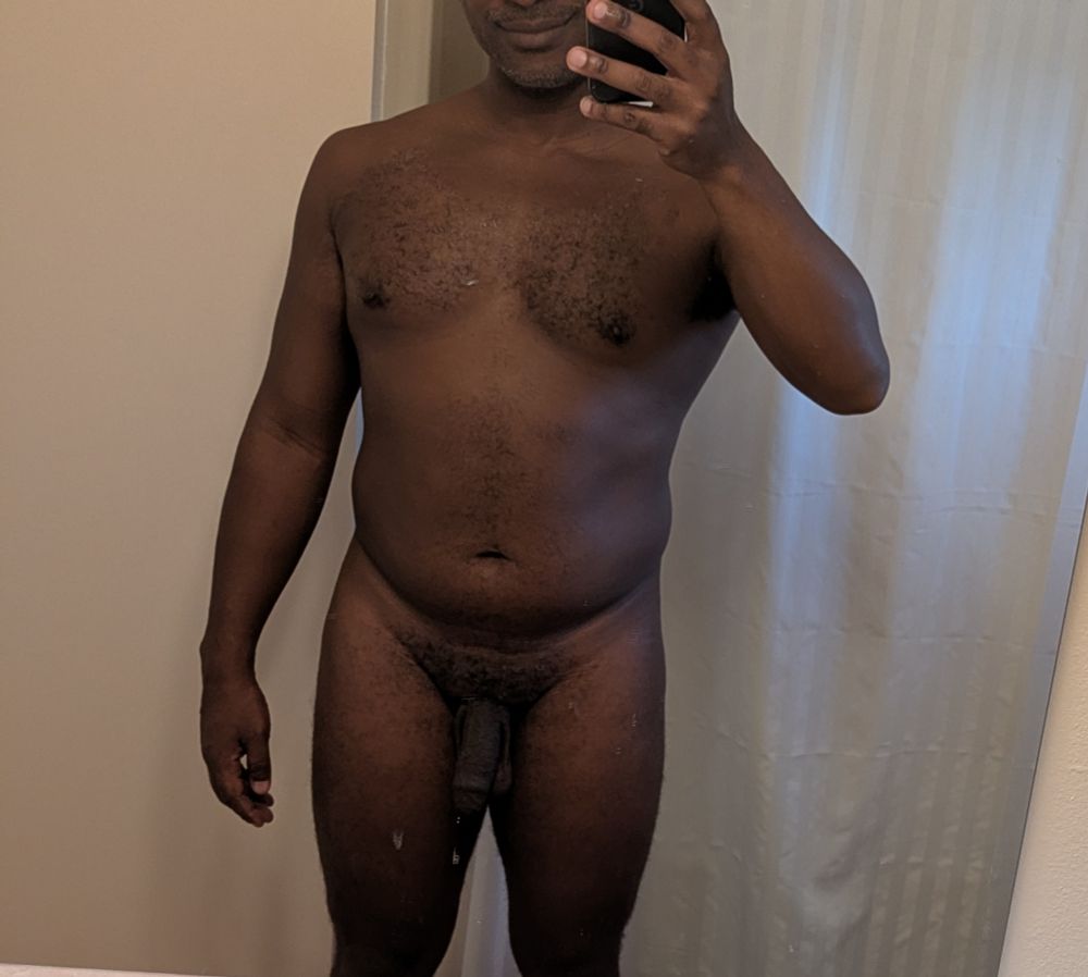 Naked man with a hairy chest takes a mirror selfie in a bathroom.