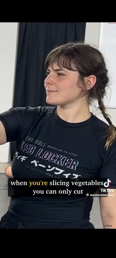 Close up of a woman in a self-defense class looking very doubtful about her instructor's facts on this one. She has dark hair in a plait to her shoulder with bangs, and is wearing black sweats and a black T-shirt.