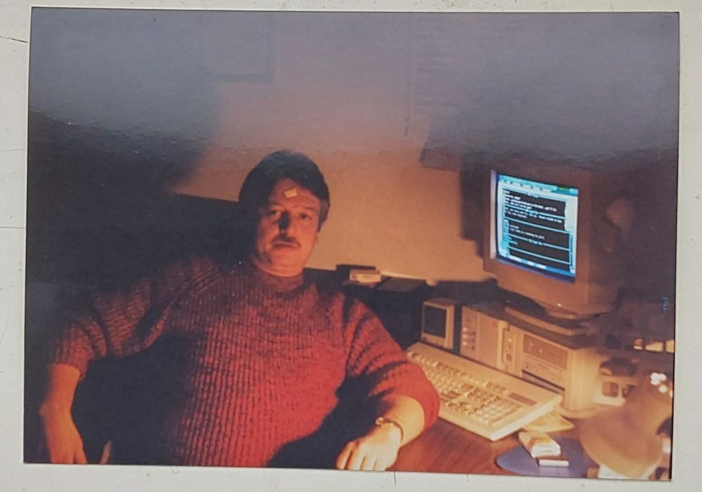 A photo (taken 2025) of a photo (from the 90s?), this is Dave, a white man in a red jumper sitting at his desk with a pc.