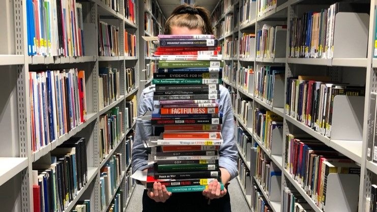 A person stood amongst the SSL book shelves carrying a pile of books in front of them. The pile is so high it is obscuring their face.