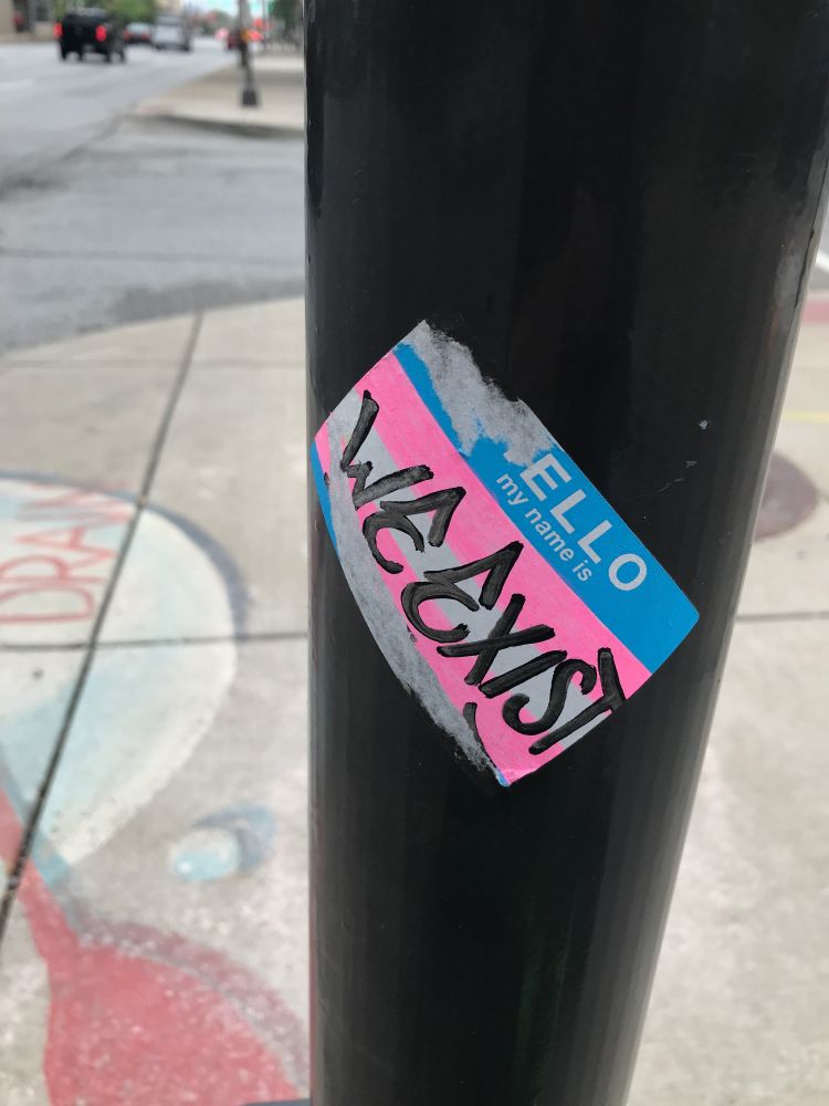 sidewalk of Fort Wayne, Indiana - name tag sticker colored as the trans flag with the text "Hello, my name is : WE EXIST"