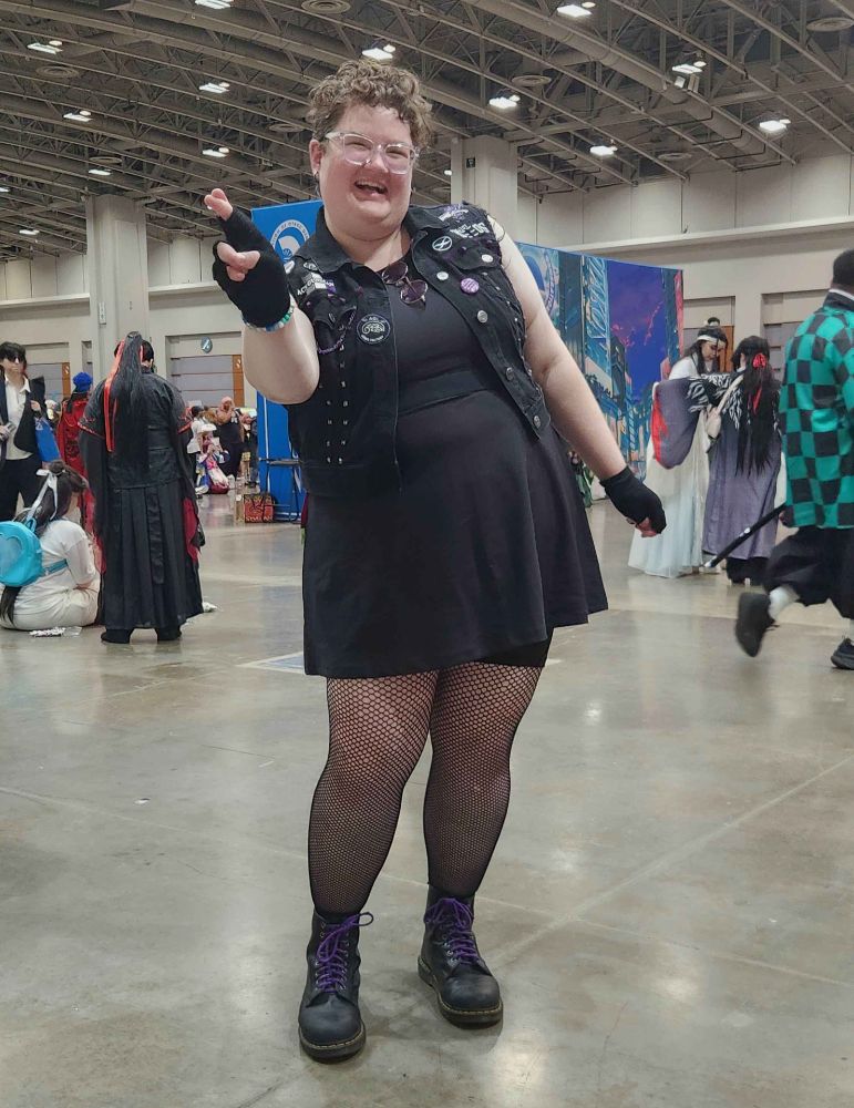 Photo of Maggie at Otakon 2024. She is wearing an outfit inspired by Vash in episode 12 of Trigun Stampede. It consists of a black dress, fishnet tights, and black combat boots with purple laces. Over the dress she is wearing a black denim vest with patches and decorations inspired by the series. More details can be seen in the other photos. Maggie is grinning at the camera with her fingers crossed, mimicking Vash's "love and peace" pose from the 1998 anime.