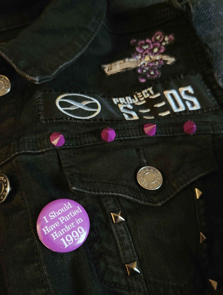 Close up photo of the vest Maggie is wearing in the first photo. This part of the vest has a Project Seeds rectangular patch, a purple button that reads "I should have partied harder in 1999," and a patch of Vash's gun surrounded by purple flowers. There are silver/gunmetal studs and purple studs decorating the vest.