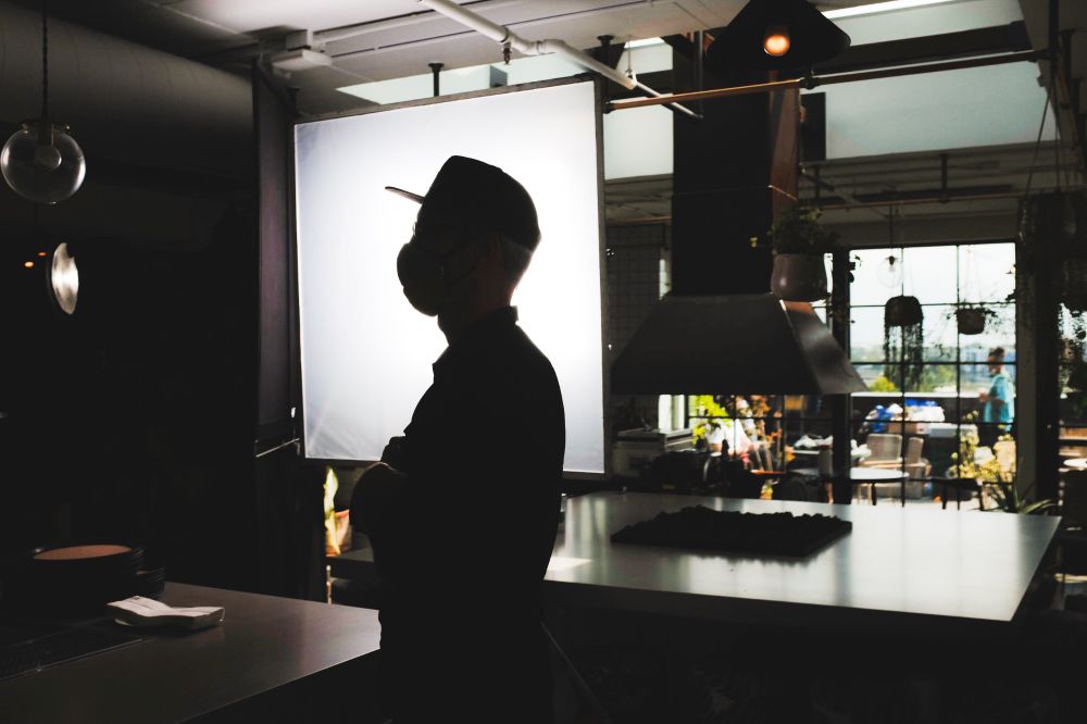 A film set during a still moment. A crew member stands in front of a 4x4 silk, which silhouettes them in the image. The space appears to be a fancy, industrial kitchen. The interior is dark, save for the light source the crew member stands in front of, and light spills onto a metal table with a stove burner from an outside window in the distance of the photo