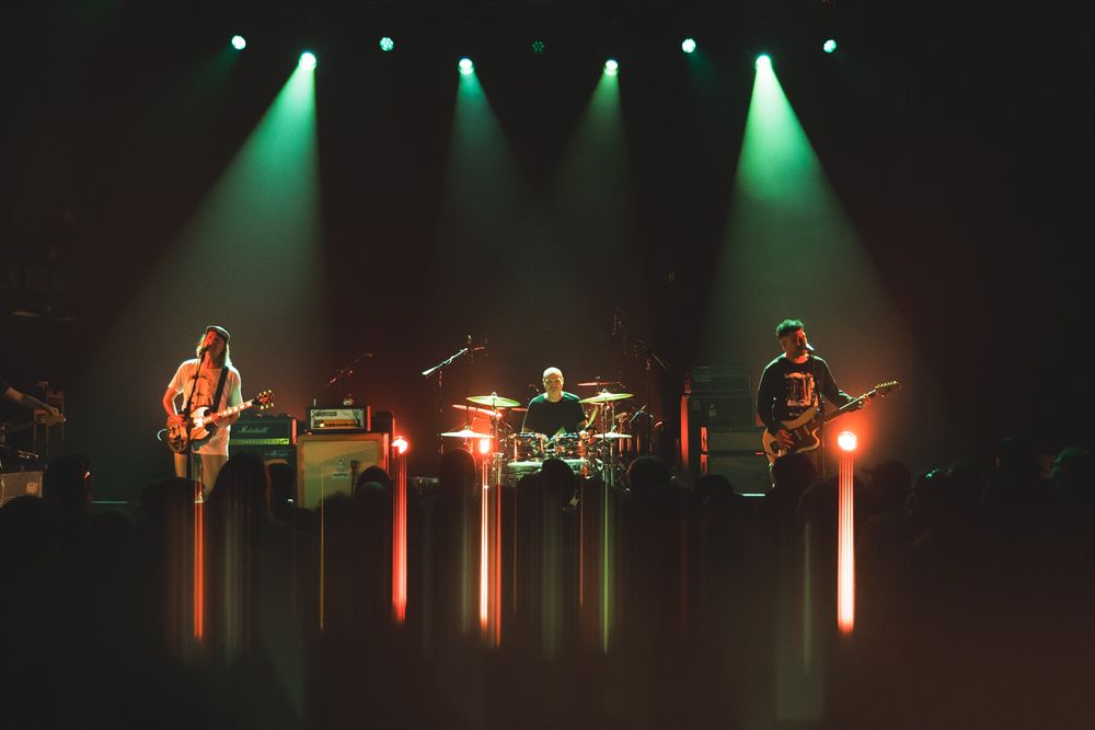 Post-hardcore band, Quicksand performs on stage. Picture is a wide shot of the stage, lights above shine a green tint while backing lights and front lights shine a reddish-orange. Below the band, light streaks fade into the crowd from a specialty lens filter. 