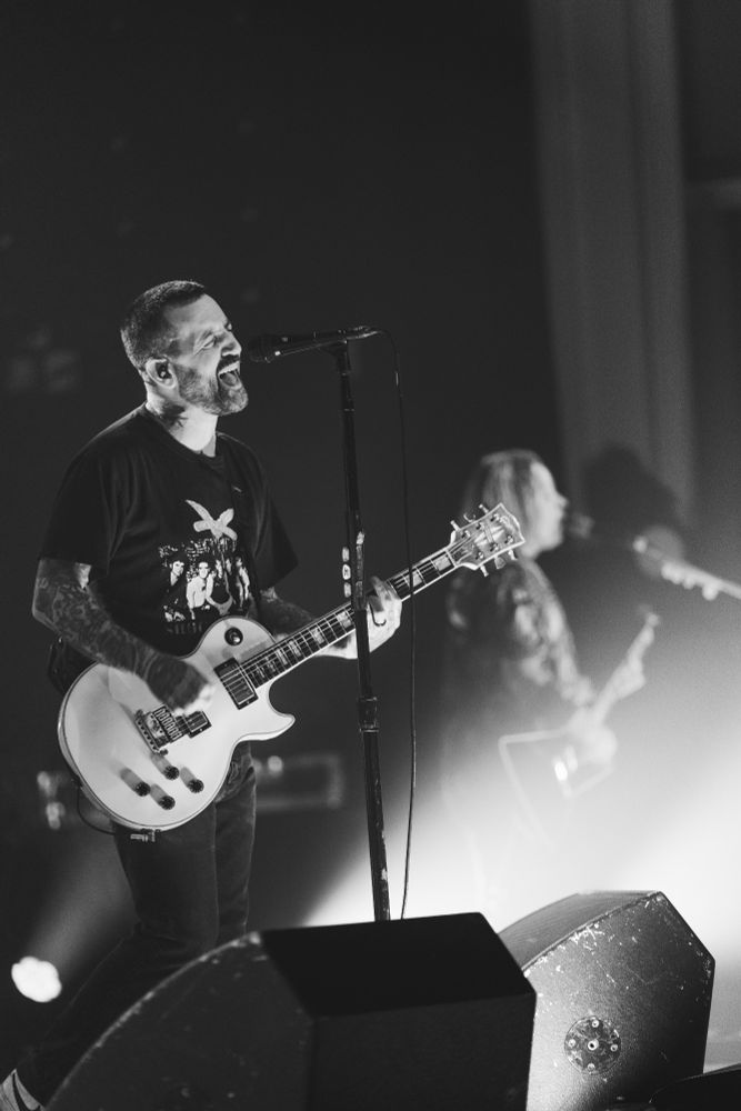 Black and white: 2 guitarists on stage, one is blurry in the background and slightly covered by light, the one in focus in the foreground is playing a white guitar and signing into a microphone