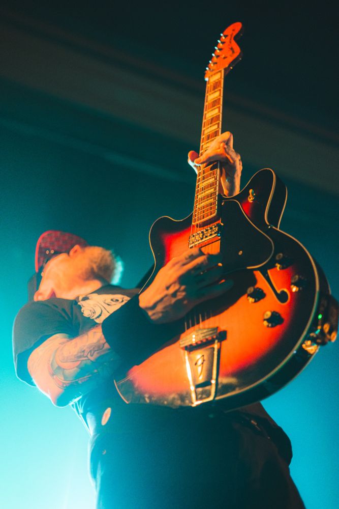 The singer and guitarist of the Smoking Popes holds his guitar high, straight up and down while playing a solo. Blue light surrounds him in the background