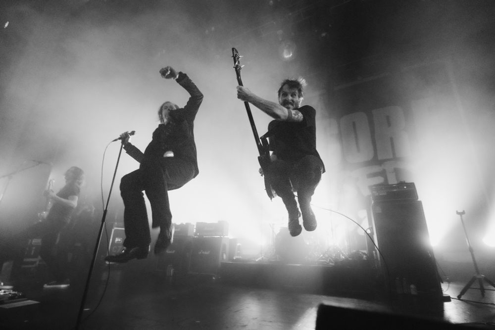 The singer and bass player of the band, Refused, airborne and captured on camera mid-jump. Bright white lights shine behind them, behind their amps and the drum set. Their guitar player is engulfed in light on the left side of the image. Image is black and white 