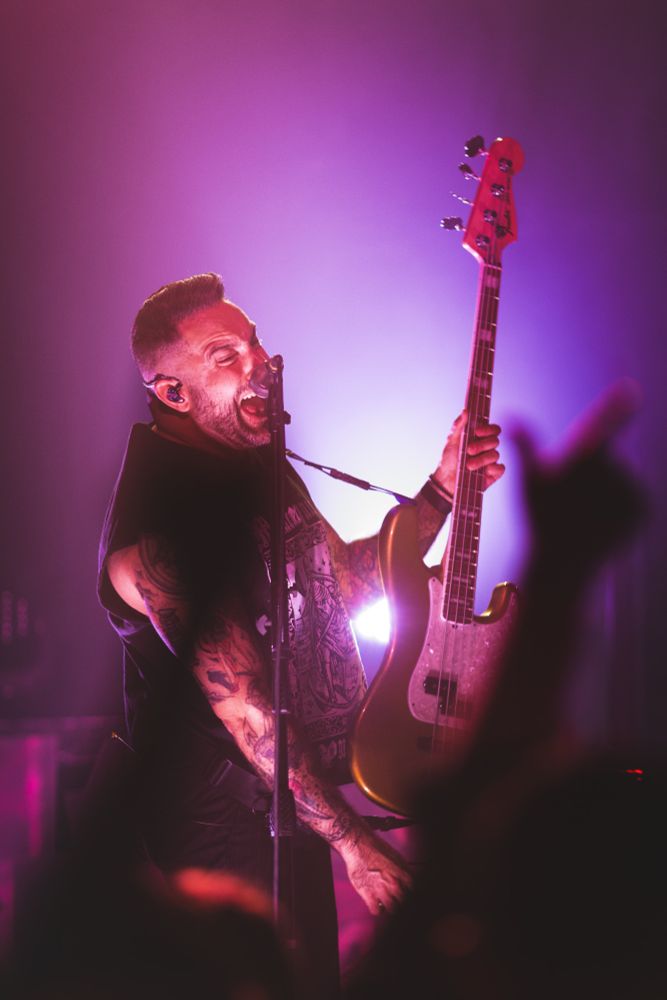 The bass player of Bayside sings into a microphone while holding his bass up. Hands are blurred in the foreground of the image. A purple, violet and pink light shines in the background
