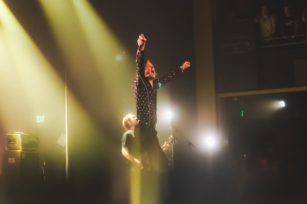The singer of Refused  stands tall with his arms out to the crowd, yelling with them as they sing along to a song. Bright yellow and white lights shine from above and behind. Haze fills the stage allowing the light to shine in beams on and behind the band. The lower 1/4 of the image is a bit of a light streak from a specialty lens filter 