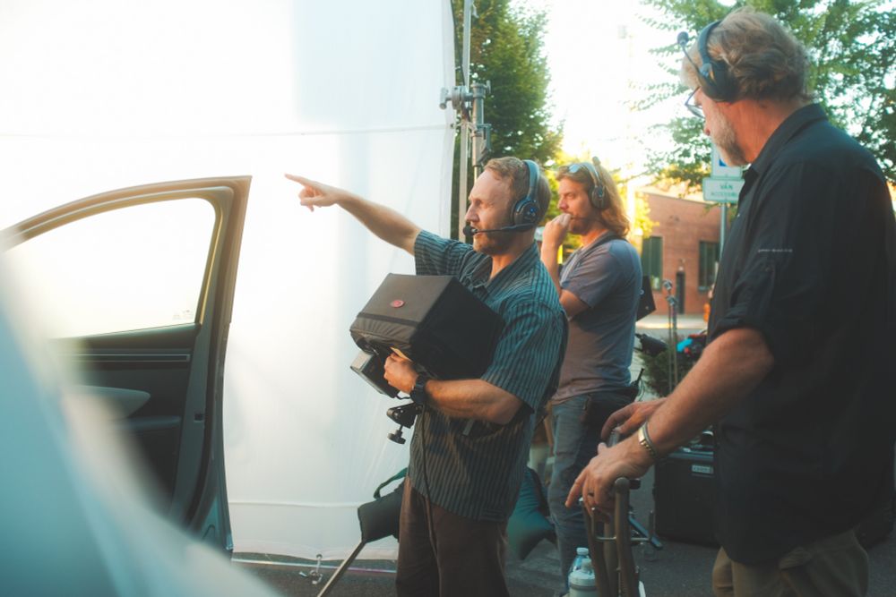 A director of photography directs the grips on how he wants the lighting. He is holding a large monitor and pointing while two other crew members listen for adjustments. A 12x12 scrim sits behind them from the middle of frame to the left