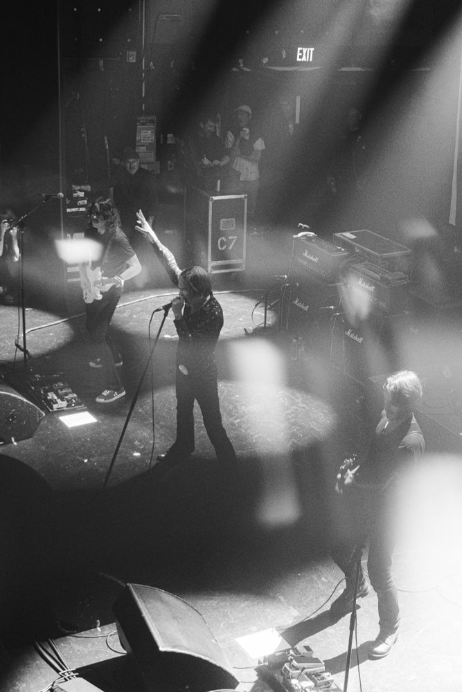 A black and white image of the band, Refused, performing on stage. POV of from a balcony above and to the right of the stage. A slight blurring effect cuts through the middle of the image from a specialty lens filter. The stage lights create islands of light and streams of darkness. 