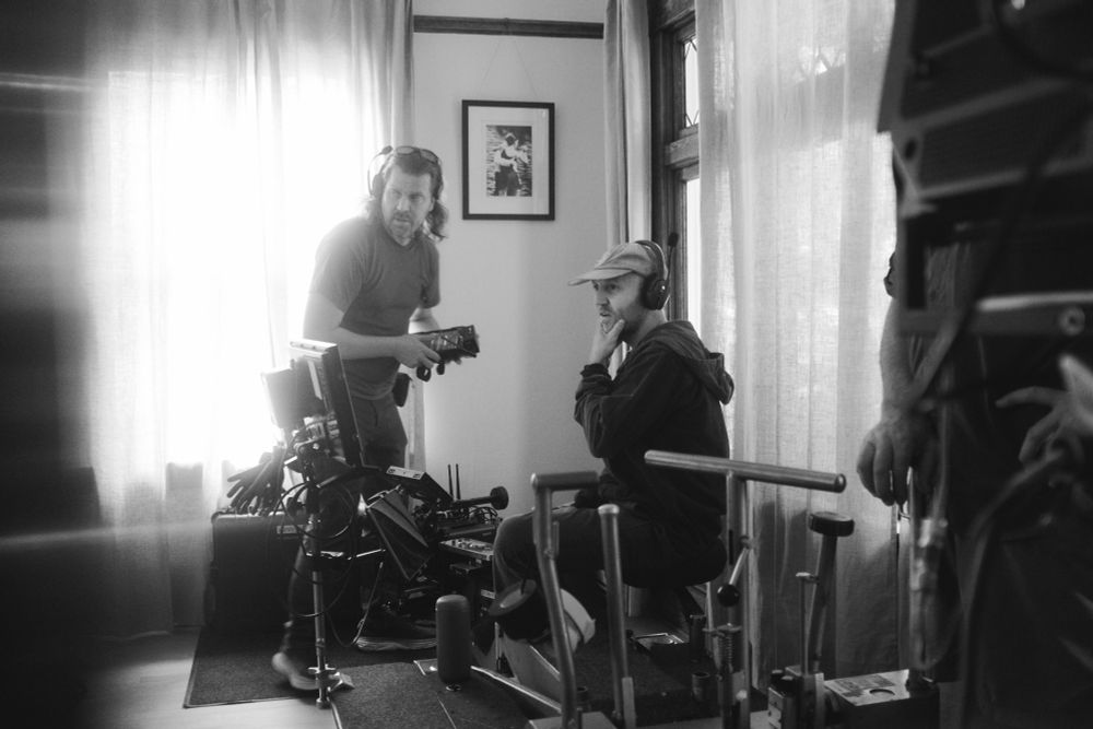 Monochrome photo of Behind the scenes on a commercial film shoot. A DP and Gaffer look at their frame on a monitor to adjust lighting. They are inside a house, with windows letting in bright ambient light from outside. 
