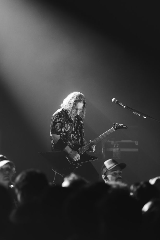 Black and white - a guitar player of the band bayside plays guitar on stage, a shaft of light illuminates him from behind, with the background fading into black. The crowd is blurry in the foreground at the bottom of the image 