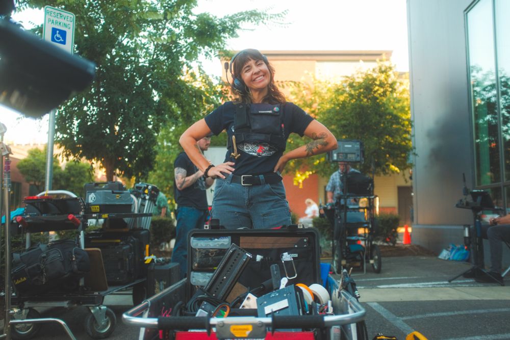 A crew member on a film shoot smiles as their picture is taken, while standing behind a cart of their camera gear