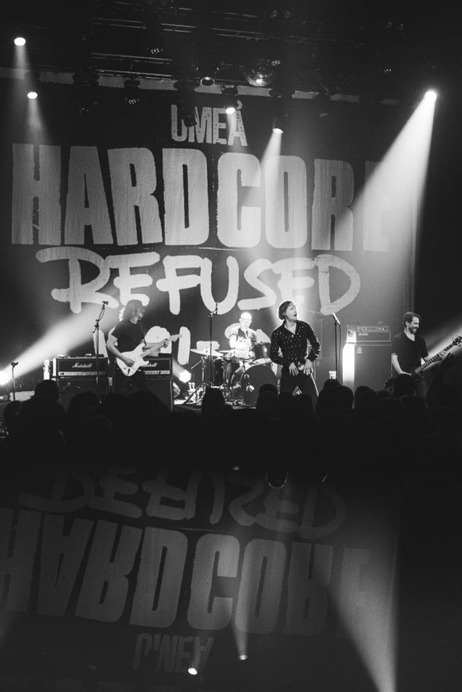 Hardcore band, Refused, performs on stage in front of a massive banner that reads “Umea Hardcore Refused.” Top half of the image is the stage from the lights down to the crowd, bottom half of the image is the crowd and a slight mirroring effect from a specialty lens filter. Image is black and white