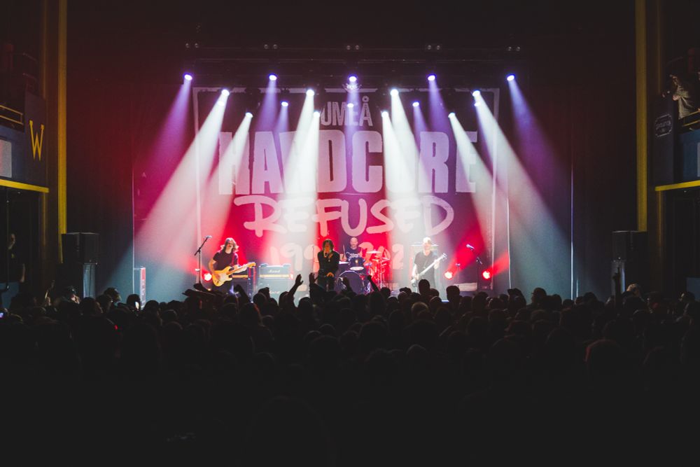 A wide picture of a band, Refused, performing on stage under bright white and purple lights. Backing lights are bright red and light up a banner that reads “Umea Hardcore Refused.” The lower third of the image is filled from left to right with a crowd of hundreds