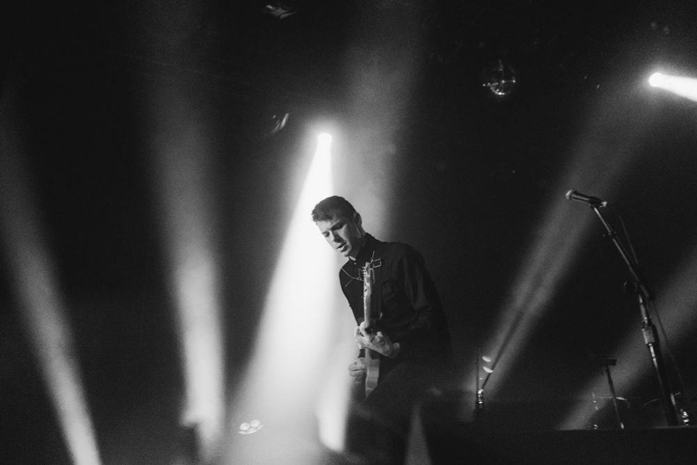 The guitarist of the band, HELP playing on stage with bright white lights behind him. Picture is monochrome