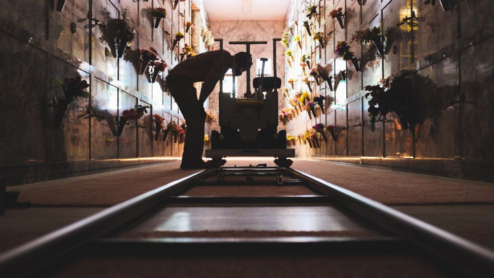 A dolly grip checks a setting on a Fischer dolly. The image sees dolly track in the foreground, and stretches into the image to where the dolly and the grip are positioned. They are inside a mausoleum. Flowers line the walls, with names of the deceased, unable to the read clearly. On the other side of the room, bright light fills a window to illuminate the space. It is on the other side of the dolly and grip and casts them in a silhouette  