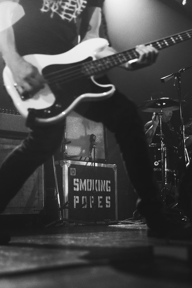Monochrome image - “smoking popes” stenciled on a guitar cabinet is in focus in the background, the bass player, cut off near the shoulder, stands closer to camera and the cabinet is framed through their legs 