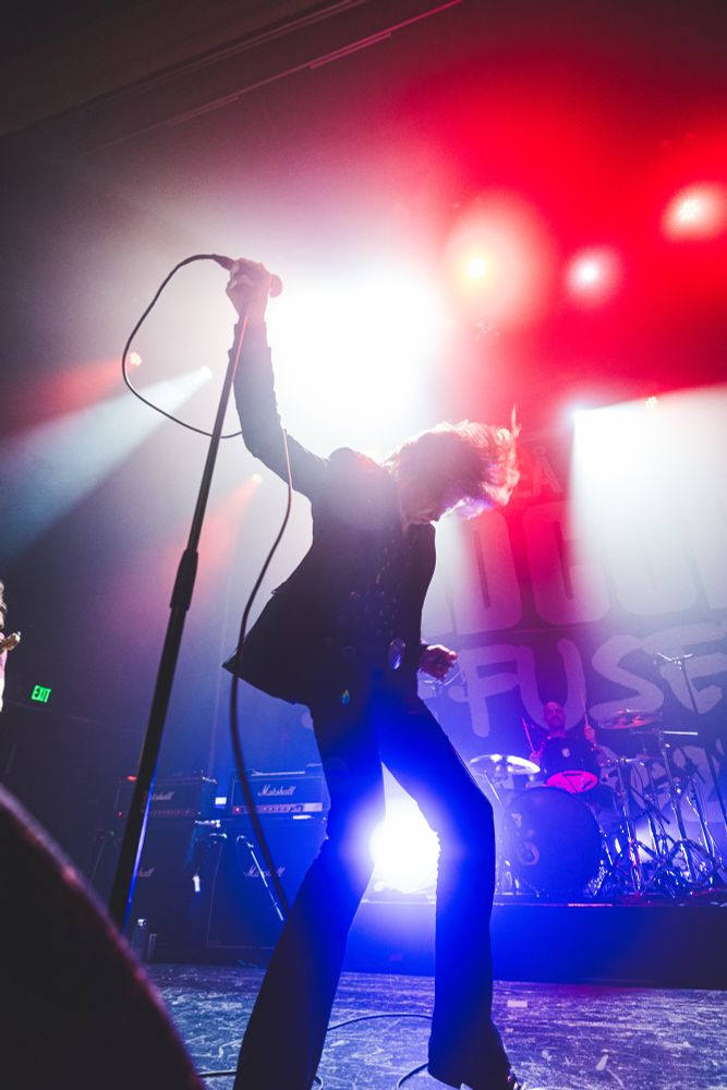 The singer of Refused holds a microphone and headbangs in front of bright lights of red, white and blue. He is nearly silhouetted by all the light 