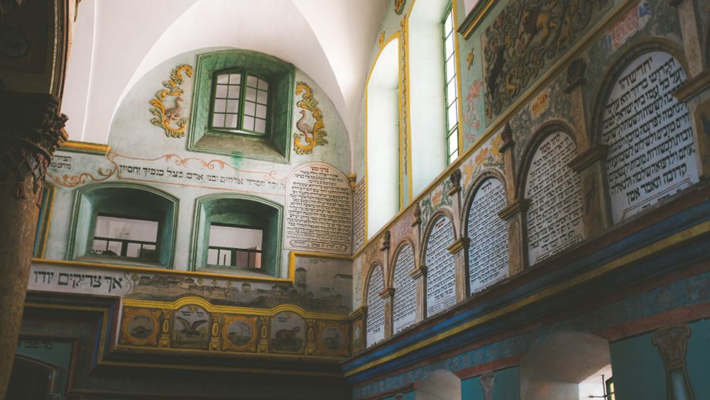 2 walls of Łańcut Synagogue, meeting in a corner near the left-center of the frame. On the right, arches with Hebrew text line the wall. Above the arches near the corner, 2 windows let in bright sunlight, illuminating the room and adjacent wall. The wall directly parallel to camera view has 3 windows high on the wall, bordered with a dark greens, intricate designs in yellow and green surround the upper most windows, another arch of Hebrew text sits beside the two lower windows and the corner where the walls meet. 