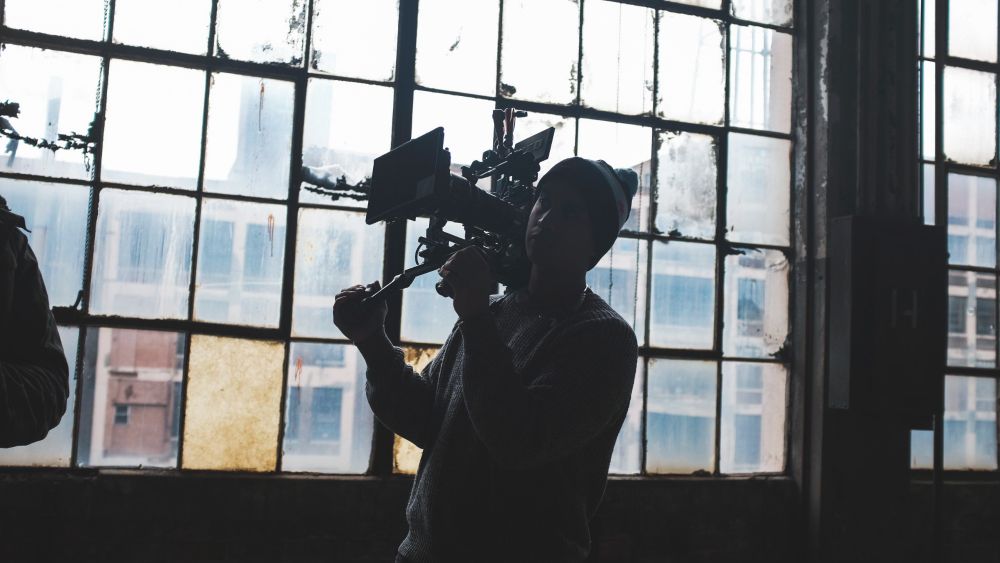 A cinematographer stands in front of a wall of old windows. The light outside is bright and casts the cinematographer in a silhouette. He holds a camera rig on his shoulder. Outside the windows, we can see another building, which has similar large panes of windows. The interior is dark and only lit from the outside