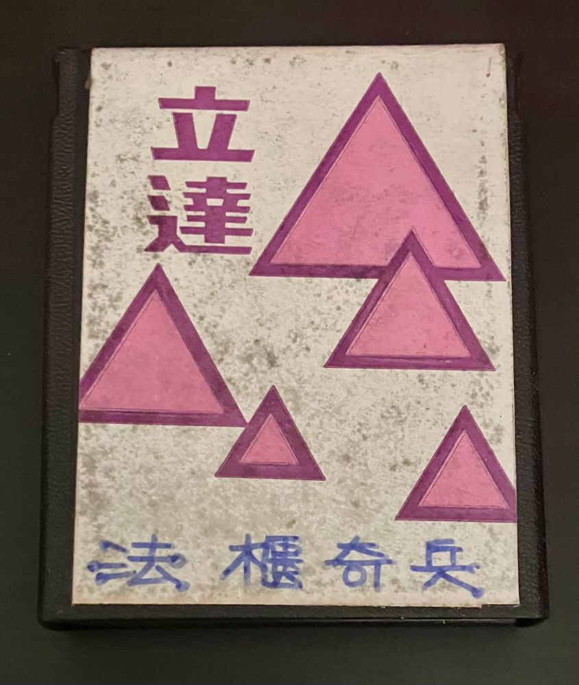An Atari 2600 cartridge with a fairly plain label. There are scattered pink triangles on a plain white background. The word 立達 (Leader?) is printed vertically in a purplish color. The name of the game has been neatly written onto the bottom of the label in blue pen. Glue spotting/staining mars the label. 