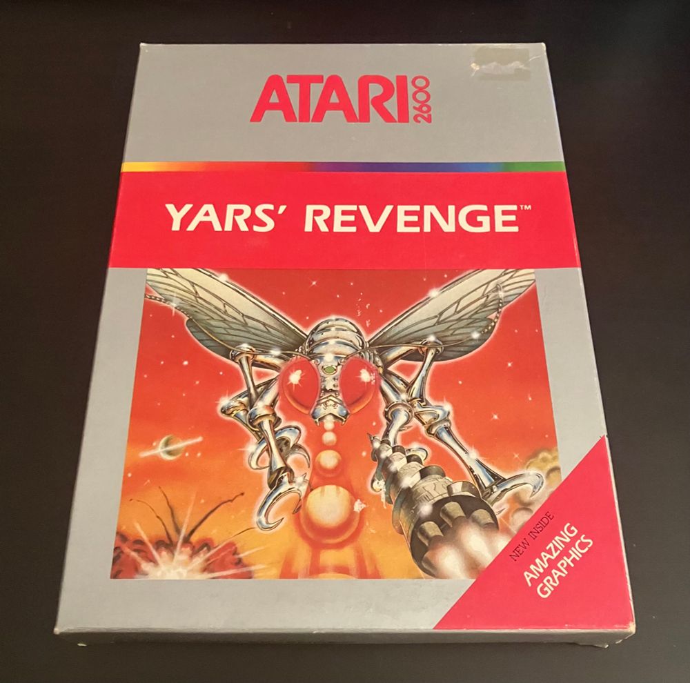 Atari 2600 box for Yars’ Revenge in the silver-label style box. Residue from a price sticker is the only major blemish. A red triangular sticker is affixed to the lower right box corner, which reads “NEW INSIDE” “AMAZING GRAPHICS”.