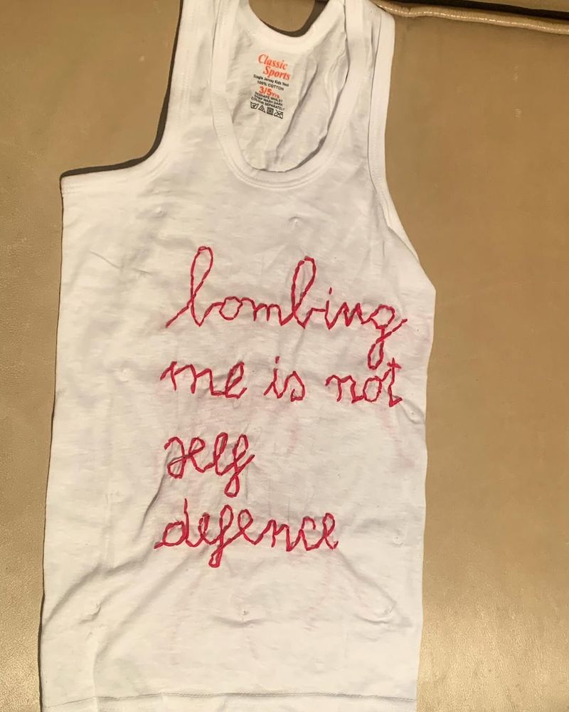 A white child's vest with the stictched words in red thread: 'Bombing me is not self defence' 