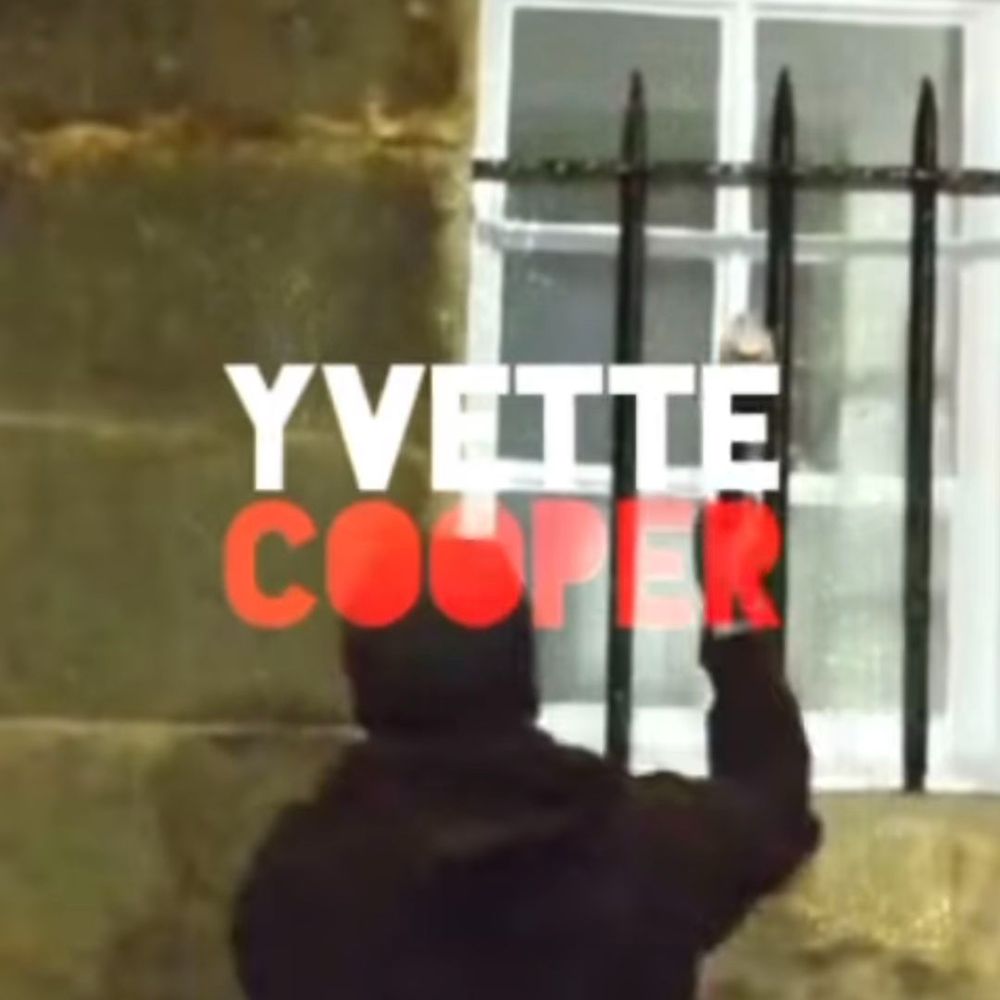 Screencap: A hooded protester smashes a window with the words 'Yvette Cooper' centred in white and red