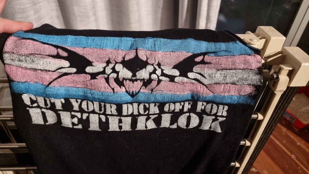 A black singlet with the trans flag painted on it. A winged skull is superimposed on the flag. The text underneath reads "CUT YOUR DICK OFF FOR DETHKLOK"