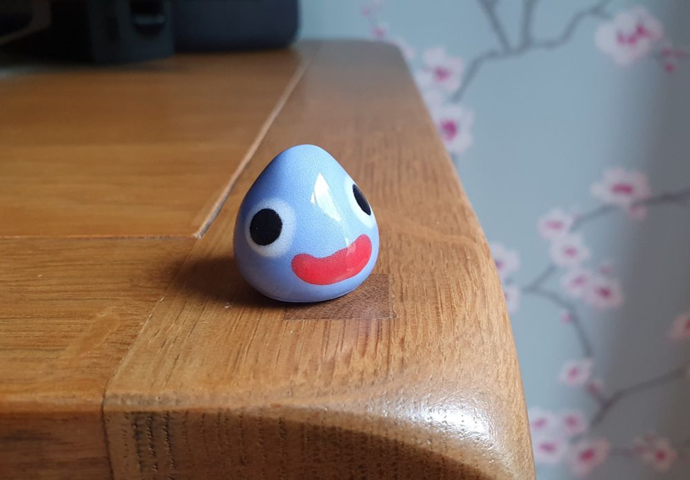 Goofy looking clay enamel Dragon Quest slime sat on the edge of a table.